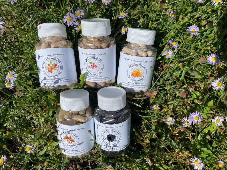 K&ouml;nnte beinhalten: F&uuml;nf transparente Plastikflaschen mit wei&szlig;en Deckeln, gef&uuml;llt mit Kapseln, beschriftet mit "Ancient Herbs Co." und verschiedenen Kr&auml;uterzutaten. Die Flaschen sind auf einem Bett aus gr&uuml;nem Gras und kleinen lila und gelben Blumen angeordnet.