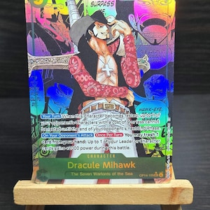 May include: A holographic trading card featuring Dracule Mihawk from "The Seven Warlords of the Sea." The card has a colorful design with the number "10000" and text that reads "STRIVE TO SURPASS." The card is displayed on a small wooden easel.