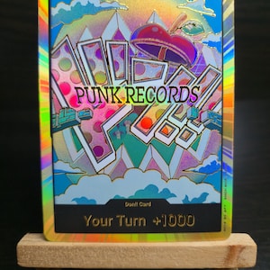 May include: A holographic trading card with a gold border, featuring the text "PUNK RECORDS" and a colorful abstract design. The card includes the text "Your Turn +1000" and is displayed on a small wooden easel.