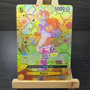 May include: A holographic trading card featuring the character Nami from the Egghead/Straw Hat Crew. The card has a gold background with a colorful illustration of Nami. The card includes text such as "5000" and "On Play".