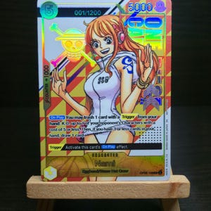 May include: A collectible trading card featuring the character Nami from the anime series One Piece. The card has a holographic design with a colorful background and text. The card is displayed on a small wooden easel.