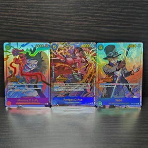 May include: Three collectible trading cards featuring anime characters. The cards display vibrant illustrations of Monkey D. Luffy, Portgas D. Ace, and Sabo. Each card has a holographic finish and includes character names and game statistics.