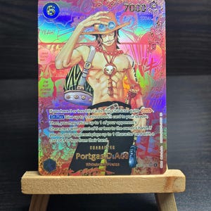 May include: A holographic trading card featuring the character Portgas D. Ace from the anime series One Piece. The card has a colorful design with the character's image, text, and game information. The card is displayed on a small wooden easel.