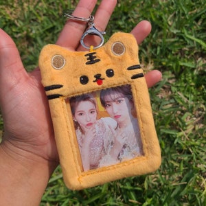 May include: A plush, yellow tiger-shaped card holder with a silver keychain. The holder features a clear window displaying two women. The tiger design includes black stripes, a black nose, and a red tongue. The background is green grass.
