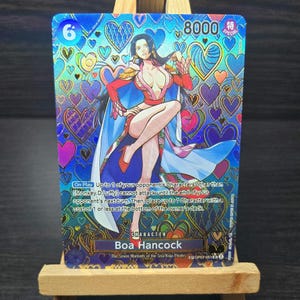 May include: A collectible trading card featuring Boa Hancock from the Seven Warlords of the Sea/Kuja Pirates. The card has a vibrant design with a character illustration, text, and a heart-themed background. The card has a value of 8000.