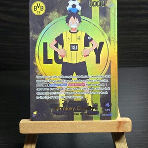 May include: A trading card featuring a character in a yellow and black soccer jersey with the text "Luffy". The card has a soccer ball on top of the character's head and the BVB logo. The card has the number 5000.