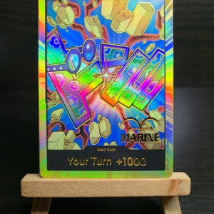 May include: A holographic trading card with a rainbow design and the word "MARINE". The card features the text "Your Turn +1000" and a colorful background with geometric shapes and a pair of glasses.