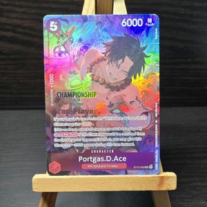 May include: A holographic trading card featuring Portgas D. Ace from the One Piece series. The card has a colorful background and the text "CHAMPIONSHIP" and "Top Player". The card also has the number "5" and "6000".