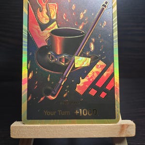May include: A gold holographic trading card with a black background featuring a black top hat with goggles, a black pipe, and flames. The text "Your Turn +1000" is printed in gold on the card.