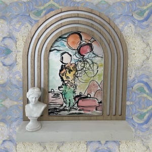 Può includere: Una cornice decorativa in legno ad arco con un dipinto ad acquerello di una figura che tiene dei palloncini. Un piccolo busto bianco di donna è su una mensola sotto il dipinto. Lo sfondo ha un motivo blu e bianco.