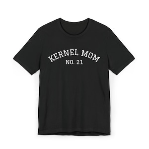 Puede incluir: Camiseta negra de cuello redondo con el texto blanco "KERNEL MOM NO. 21" impreso en el pecho. El texto está en una fuente en negrita y arqueada. La camiseta está hecha de un material suave.