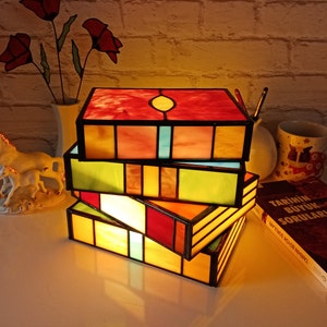 May include: A multi-layered stained glass lamp with a warm, yellow glow. The lamp is made of four stacked boxes with different colored glass panels. The lamp is sitting on a white surface with a book and a figurine.