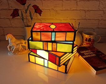 May include: A multi-layered stained glass lamp with a warm, yellow glow. The lamp is made of four stacked boxes with different coloured glass panels. The lamp is sitting on a white surface with a book and a figurine.