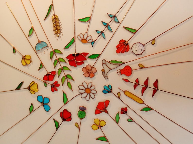 May include: A collection of colorful, stained-glass flowers and foliage, including daisies, poppies, and wheat, with copper wire stems radiating from a central point. The flowers are in shades of red, yellow, blue, and white.