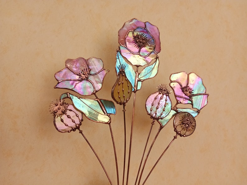 K&ouml;nnte beinhalten: Eine dekorative Glasblumenanordnung mit Kupferstielen. Die Blumen zeigen Schattierungen von Rosa, Lila und Gr&uuml;n mit irisierenden Eigenschaften. Das Design umfasst detaillierte Samenkapseln und Bl&uuml;tenbl&auml;tter.