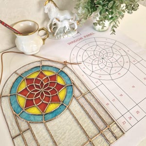 Könnte beinhalten: Ein Buntglasfenster mit einem farbenfrohen Design. Das Panel hat ein zentrales Sternmuster in Rot, Gelb und Orange, umgeben von einem blauen Kreis. Der Text "WINDOW PANEL" ist auf dem Papier neben dem Panel zu sehen.