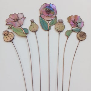 K&ouml;nnte beinhalten: Eine Sammlung handgefertigter Gartenstecker aus Glas in Form von Blumen. Die Blumen haben irisierende rosa, lila und gr&uuml;ne Glasbl&auml;tter und -bl&auml;tter mit Kupferdrahtstielen. Die Kollektion umfasst offene Bl&uuml;ten und Samenkapseln.