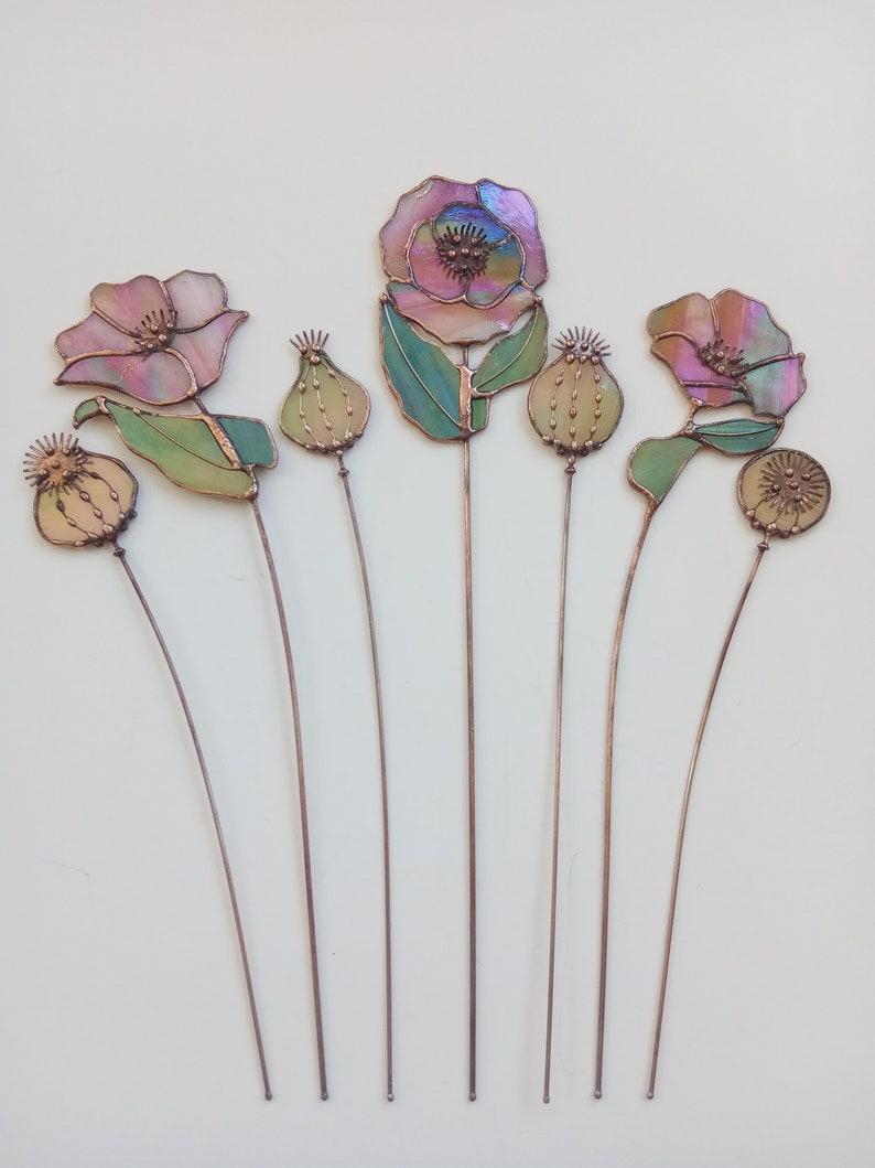 K&ouml;nnte beinhalten: Eine Sammlung handgefertigter Gartenstecker aus Glas in Form von Blumen. Die Blumen haben irisierende rosa, lila und gr&uuml;ne Glasbl&auml;tter und -bl&auml;tter mit Kupferdrahtstielen. Die Kollektion umfasst offene Bl&uuml;ten und Samenkapseln.