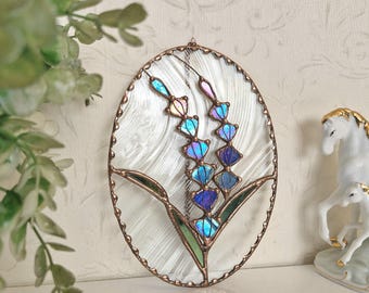 May include: An oval stained glass artwork depicting a lavender flower. The glass has a textured white background, with iridescent blue, purple, and green petals. The piece is framed with a copper-coloured metal.