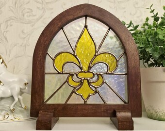 May include: A stained glass window featuring a yellow fleur-de-lis design within a dark brown wooden frame. The arched window is supported by a wooden base, and the glass is clear.