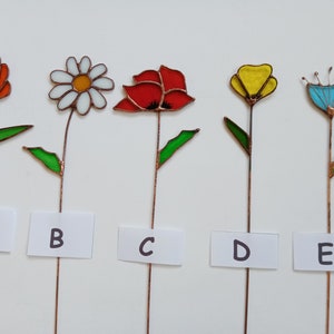 May include: Five colorful stained glass flower garden stakes. Each flower has a different design and color, including orange, white, red, yellow, and blue. The flowers are mounted on copper stems with green leaves. Each stake has a white label with a letter.
