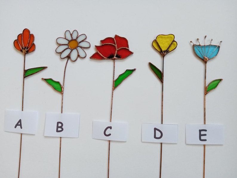 May include: Five colorful stained glass flower garden stakes. Each flower has a different design and color, including orange, white, red, yellow, and blue. The flowers are mounted on copper stems with green leaves. Each stake has a white label with a letter.