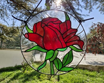 May include: A circular stained glass window depicting a red rose with green leaves and stem. The design is set against a clear, textured background and is suspended by black cords. A decorative piece for the home.