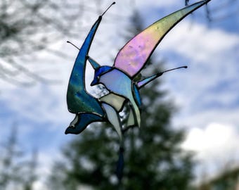 May include: A stained glass butterfly in shades of blue, green, and purple, with iridescent wings. The butterfly has a detailed design and is suspended against a blurred background of trees and sky.
