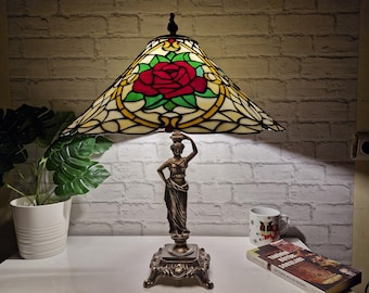 May include: A stained glass table lamp with a red rose design on the shade. The lamp has a bronze base with a classical female figure. The lamp is on a white surface with a plant and a book nearby.