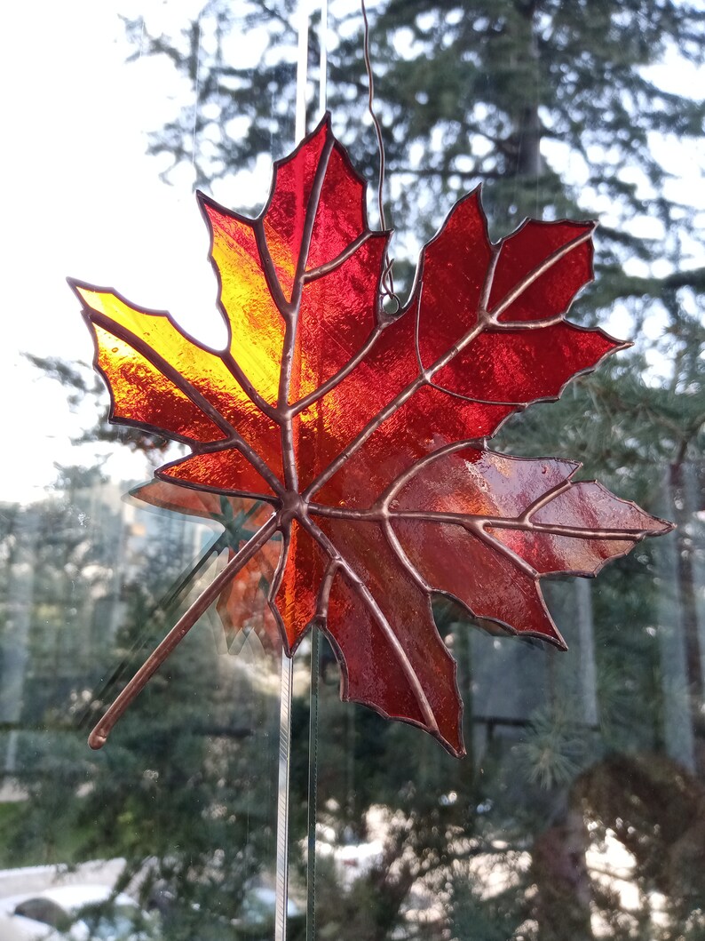May include: A stained glass maple leaf with red and orange hues. The leaf is hanging in a window with a blurred background of trees.