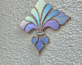 May include: A stained glass fleur-de-lis in shades of blue, purple, and green, with copper-coloured edging. A clear crystal prism hangs from the bottom. The design is mounted on a textured white wall.