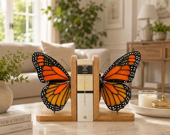 May include: A pair of wooden bookends featuring stained glass monarch butterfly wings in vibrant orange, yellow, and black. The wings are attached to light-toned wooden bases, holding a book with the text Hasan Ali Yucel and Sefiller-II.