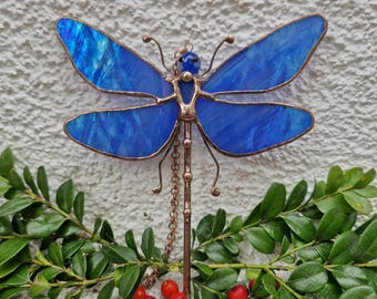 May include: A stained glass dragonfly with blue wings and a copper frame. The dragonfly has a beaded head and a chain extending from its body. The image is set against a white background with green leaves and red berries.