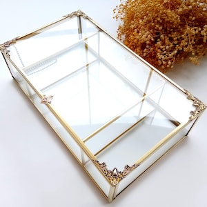 May include: A rectangular glass and gold-coloured metal display box. The box has a hinged lid and decorative corner accents. The interior is divided into sections. Dried flowers are in the background.