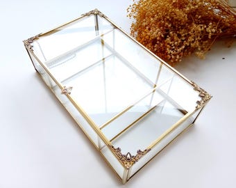 May include: A rectangular glass and gold-coloured metal display box. The box has a hinged lid and decorative corner accents. The interior is divided into sections. Dried flowers are in the background.