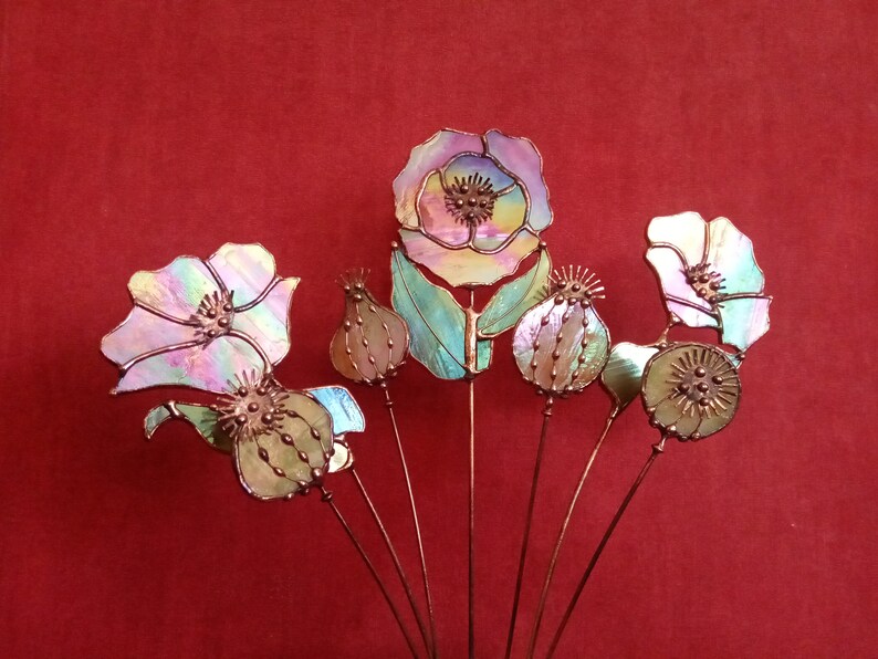 K&ouml;nnte beinhalten: Eine Sammlung von irisierenden Glasblumenskulpturen mit Kupferstielen. Die Blumen umfassen Mohnblumen und Samenkapseln, mit einer Farbpalette von Rosa, Gr&uuml;n und Gelb. Der Hintergrund ist einfarbig rot.