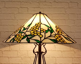 May include: A stained glass lamp with a cream shade, decorated with yellow floral clusters and green leaves. The lamp has a dark brown metal base and a geometric design. The lamp is lit, giving off a warm light.