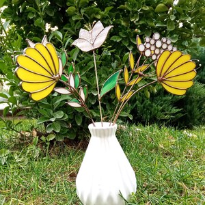 以下が含まれることがあります： テクスチャ加工された表面を持つ白いセラミックの花瓶に、ステンドグラスの花束が飾られています。花は黄色、白、茶色です。花瓶は緑の芝生の上に置かれています。