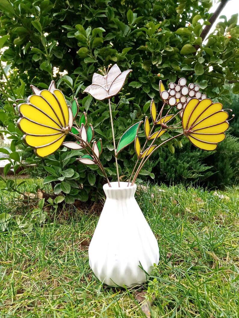 以下が含まれることがあります： テクスチャ加工された表面を持つ白いセラミックの花瓶に、ステンドグラスの花束が飾られています。花は黄色、白、茶色です。花瓶は緑の芝生の上に置かれています。
