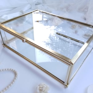 May include: A rectangular glass and gold-coloured metal box with a hinged lid. The box has a minimalist design with clean lines. The clear glass allows visibility of the contents. Decorative elements such as pearls and flowers surround the box.