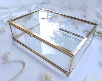 May include: A rectangular glass and gold-coloured metal box with a hinged lid. The box has a minimalist design with clean lines. The clear glass allows visibility of the contents. Decorative elements such as pearls and flowers surround the box.