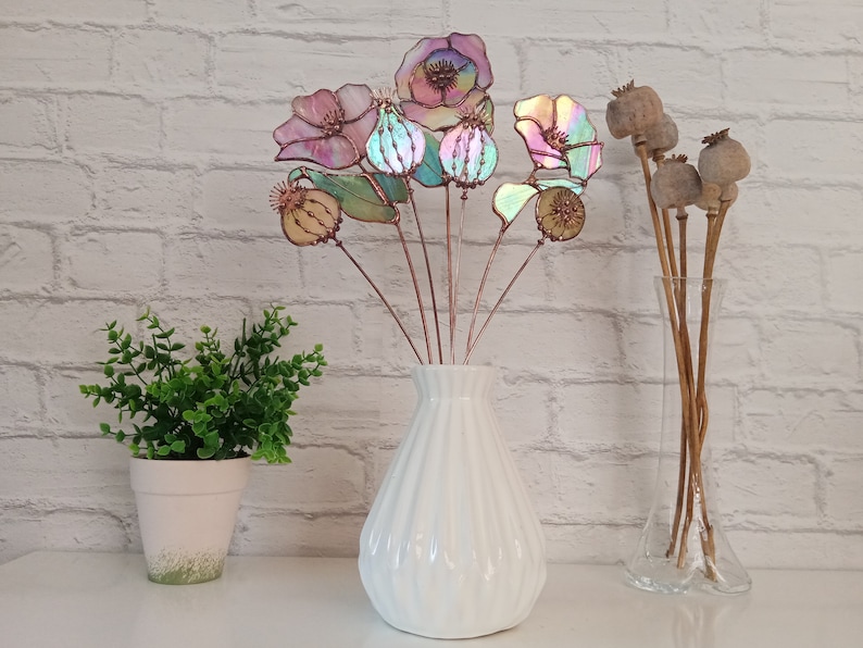 K&ouml;nnte beinhalten: Eine wei&szlig;e Vase enth&auml;lt einen Strau&szlig; aus irisierenden Glasblumen mit Kupferstielen. Die Blumen haben rosa, blaue und gr&uuml;ne Farbt&ouml;ne. Getrocknete Samenkapseln befinden sich in einer durchsichtigen Vase.
