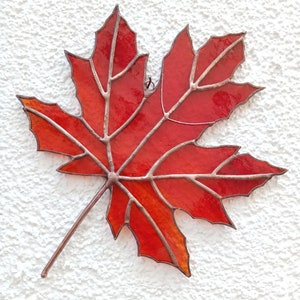 May include: A stained glass maple leaf with a copper frame. The leaf is red and orange with a gradient of color. The leaf is hanging on a white wall.