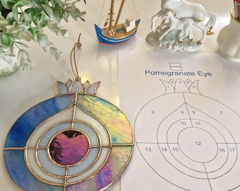 May include: A colourful stained glass pomegranate decoration with blue, white, and iridescent panels. The ornament has a copper-coloured frame and is suspended by a thin gold string. A diagram labelled "Pomegranate Eye" is next to the ornament.
