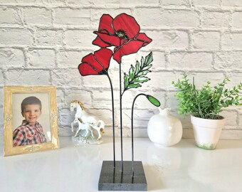 May include: A stained glass poppy sculpture featuring two red flowers and green leaves on black stems, set on a dark grey square base. A framed photo, a white horse figurine, and a potted plant are in the background.
