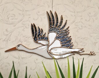 May include: Stained glass crane in flight, with white, black, and blue glass and copper edging. The bird's wings are spread, and the beak is orange. A decorative piece for home decor.