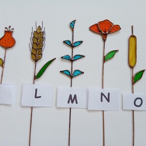 May include: Five colorful stained glass plant stakes with copper stems. The designs include a flower, wheat, a leafy plant, an orange flower, and a cattail. Each stake has a white label with a letter: K, L, M, N, and O.