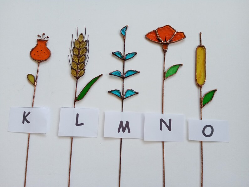 May include: Five colorful stained glass plant stakes with copper stems. The designs include a flower, wheat, a leafy plant, an orange flower, and a cattail. Each stake has a white label with a letter: K, L, M, N, and O.