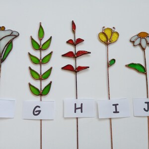 May include: Five stained glass flower garden stakes. The flowers include a white calla lily, green leaves, red leaves, a yellow flower, and a white daisy. Each flower is mounted on a copper wire stem. The letters F, G, H, I, and J are on white labels.