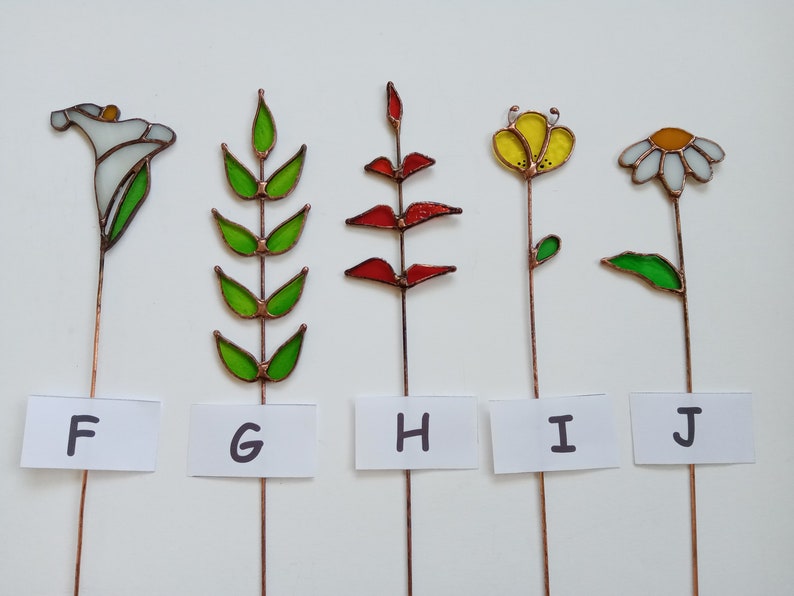 May include: Five stained glass flower garden stakes. The flowers include a white calla lily, green leaves, red leaves, a yellow flower, and a white daisy. Each flower is mounted on a copper wire stem. The letters F, G, H, I, and J are on white labels.